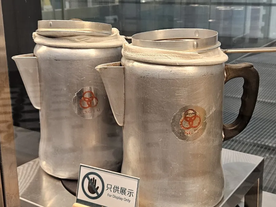 兩個鋁製奶茶壺，套著白布袋，壺身標有三個紅圈和 TING TAI 字樣，前面放著「只供展示」牌子。